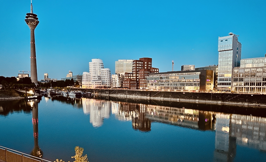 Ein abendlicher Blick auf den Düsseldorfer MedienHafen, bei dem der Rheinturm und moderne Architektur sich im ruhigen Wasser spiegeln.