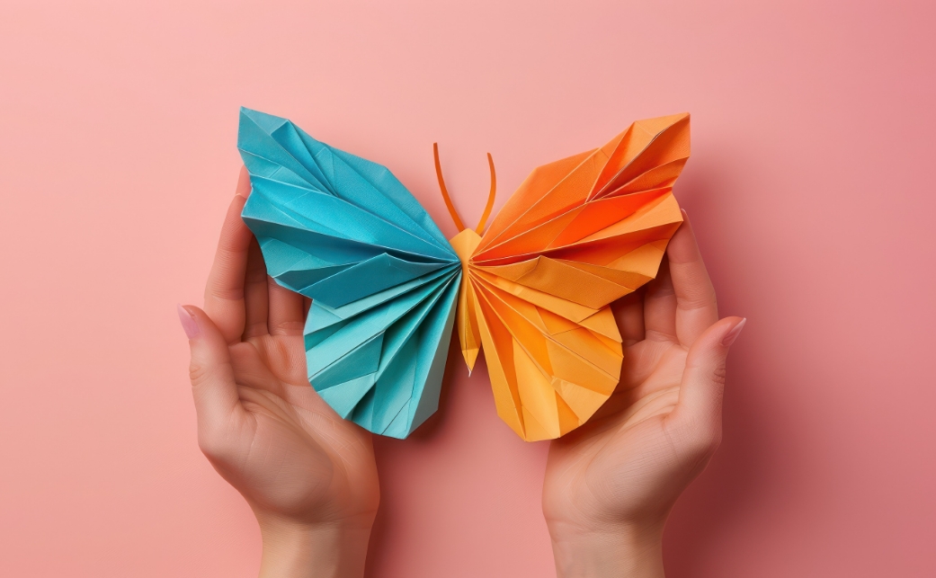 Ein Origami-Schmetterling in Blau und Orange wird sanft in zwei Händen gehalten.